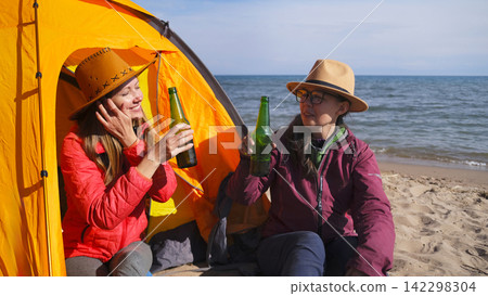 Two happy women in love are sitting on the beach and relaxing with beer bottles in their hands. Young female students smile Two happy women in love are sitting on the beach and relaxing with beer bottles in their hands. Young female students smile 142298304