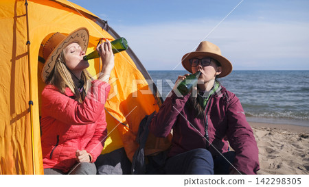 Two tourists drink beer at a tent on the beach. Two tired travelers are relaxing by the sea. Freedom, relaxation and unity with nature Two tourists drink beer at a tent on the beach. Two tired travelers are relaxing by the sea. Freedom, relaxation and unity with nature 142298305