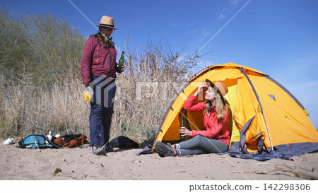 Two girlfriends are relaxing on the beach by the tent. Women eat snacks and drink beer outdoors. Female alcoholism. Tourism, trekking and adventures Two girlfriends are relaxing on the beach by the tent. Women eat snacks and drink beer outdoors. Female alcoholism. Tourism, trekking and adventures 142298306