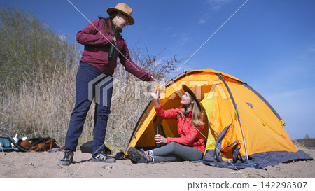 One woman hands the other some chips. Two best friends are drinking beer and eating snacks outdoors. Tourists relax on the beach with a tent One woman hands the other some chips. Two best friends are drinking beer and eating snacks outdoors. Tourists relax on the beach with a tent 142298307