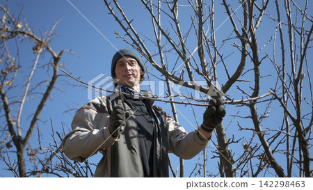 A happy young amateur gardener is working in the garden. A man in a hat and gloves holds a pruner in his hands and prepares to cut the dry branches of a tree A happy young amateur gardener is working in the garden. A man in a hat and gloves holds a pruner in his hands and prepares to cut the dry branches of a tree 142298463
