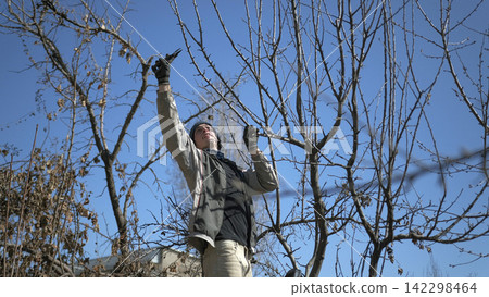 A young specialist in pruning dry branches from garden trees. A man prunes a tree with garden scissors. He is wearing a black jacket, hat and gloves A young specialist in pruning dry branches from garden trees. A man prunes a tree with garden scissors. He is wearing a black jacket, hat and gloves 142298464