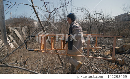 A construction worker carries a ladder in a wooded area. A man is working in an abandoned garden. Pruning old trees. Part-time job for students 142298469