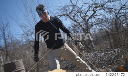 A man is chopping firewood. The lumberjack. A young strong guy in a black shirt and a blue bandana holds an axe and a log in his hands. The action takes place in a wooded area A man is chopping firewood. The lumberjack. A young strong guy in a black shirt and a blue bandana holds an axe and a log in his hands. The action takes place in a wooded area 142298472