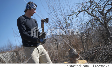 Harvesting of firewood. A handsome man is standing in a field with an axe in his hands and chopping firewood. He is wearing a black shirt and beige trousers Harvesting of firewood. A handsome man is standing in a field with an axe in his hands and chopping firewood. He is wearing a black shirt and beige trousers 142298473