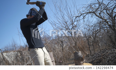 A man is chopping firewood in an abandoned garden. A guy is swinging an axe in a wooded area. The man is wearing a bandana and a black shirt A man is chopping firewood in an abandoned garden. A guy is swinging an axe in a wooded area. The man is wearing a bandana and a black shirt 142298474
