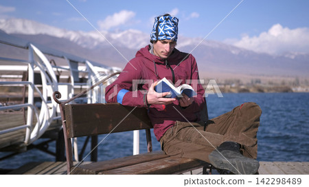 A lonely, handsome student sits on a bench by the water and reads a book against the backdrop of the Kyrgyz mountains. Lake Issyk Kul is the pearl of Central Asia A lonely, handsome student sits on a bench by the water and reads a book against the backdrop of the Kyrgyz mountains. Lake Issyk Kul is the pearl of Central Asia 142298489