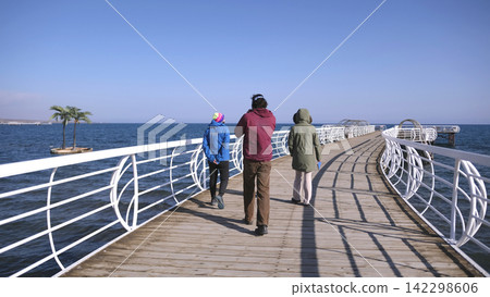 Issyk Kul Lake boardwalk in Kyrgyzstan. Three friends are walking along the pier on a sunny day. The scene is peaceful and relaxing Issyk Kul Lake boardwalk in Kyrgyzstan. Three friends are walking along the pier on a sunny day. The scene is peaceful and relaxing 142298606