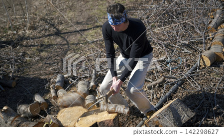 A farmer in a blue bandana is chopping firewood on a garden plot on a sunny, clear day. The man is wearing a black shirt and khaki trousers A farmer in a blue bandana is chopping firewood on a garden plot on a sunny, clear day. The man is wearing a black shirt and khaki trousers 142298627