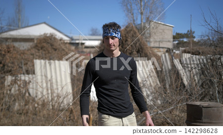A handsome young man with a bandana on his head stands in front of a stack of firewood. He holds an axe in his hand. A village guy is working A handsome young man with a bandana on his head stands in front of a stack of firewood. He holds an axe in his hand. A village guy is working 142298628
