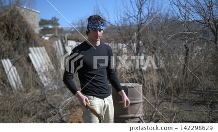 Spring work at the cottage. Garden cleaning. Chopping firewood. A man in a black shirt and a blue bandana holds an axe in his hands Spring work at the cottage. Garden cleaning. Chopping firewood. A man in a black shirt and a blue bandana holds an axe in his hands 142298629