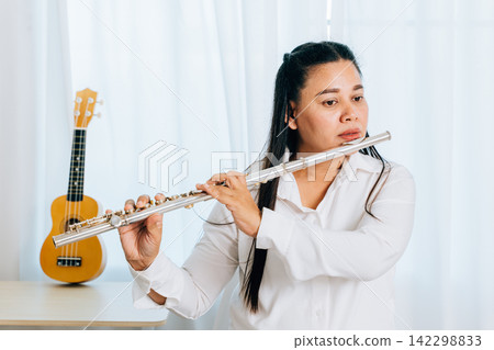 A flutist in a white shirt sits on a chair at home, blowing into her silver flute. The living room curtain backdrop accentuates 142298833