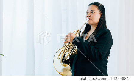 Dressed in black, a woman blows a gleaming French horn in gold 142298844