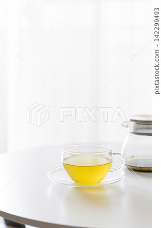 A clear glass teapot and a glass of warm green tea/Japanese tea are placed on a white table. A clear glass teapot and a glass of warm green tea/Japanese tea are placed on a white table. 142299493