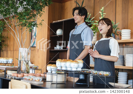 一名二十多歲的男員工和一名二十多歲的女員工面帶微笑,鼓掌,在咖啡櫃檯後面為顧客服務。 一名二十多歲的男員工和一名二十多歲的女員工面帶微笑,鼓掌,在咖啡櫃檯後面為顧客服務。 142300148