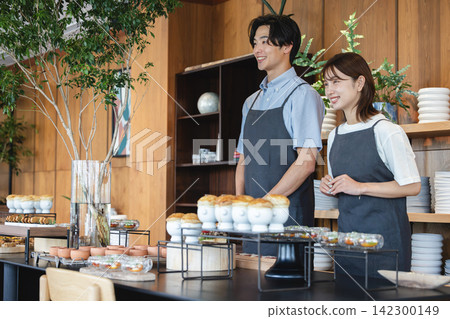 一名二十多歲的男員工和一名二十多歲的女員工面帶微笑,熱情地在咖啡櫃檯為顧客服務。 一名二十多歲的男員工和一名二十多歲的女員工面帶微笑,熱情地在咖啡櫃檯為顧客服務。 142300149