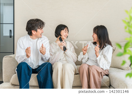 一家人悠閒地坐在客廳沙發上，手裡拿著電視遙控器。 142301146