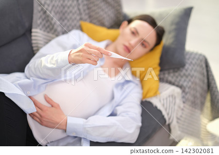 Pregnant woman lies on soft sofa holding belly and thermometer Pregnant woman lies on soft sofa holding belly and thermometer 142302081
