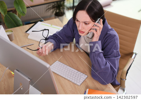 Concerned woman talks on phone while working at office computer Concerned woman talks on phone while working at office computer 142302597
