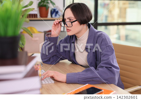 Woman adjusts glasses while focusing on computer screen at work Woman adjusts glasses while focusing on computer screen at work 142302601
