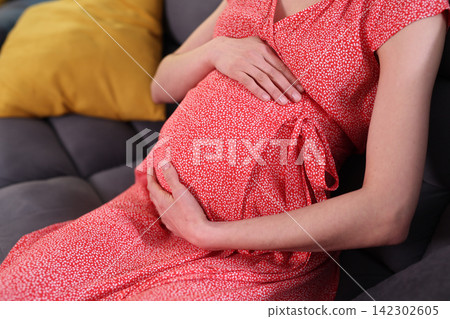 Pregnant woman gently holds belly while resting on home sofa Pregnant woman gently holds belly while resting on home sofa 142302605