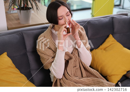 Sick woman uses nasal spray while recovering on home sofa Sick woman uses nasal spray while recovering on home sofa 142302716