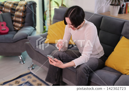 Woman checks smartphone while sitting on comfortable home sofa Woman checks smartphone while sitting on comfortable home sofa 142302723
