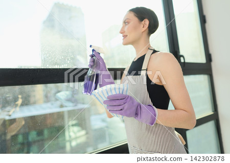 Happy maid in apron sprays detergent to clean window glass Happy maid in apron sprays detergent to clean window glass 142302878