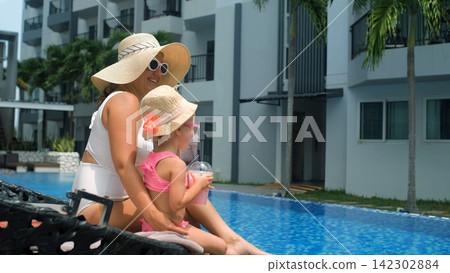 Side view of mother and little daughter both in straw hats and sunglasses drinking fruit cocktails together in pool lounge chairs. Twinning style concept Side view of mother and little daughter both in straw hats and sunglasses drinking fruit cocktails together in pool lounge chairs. Twinning style concept 142302884