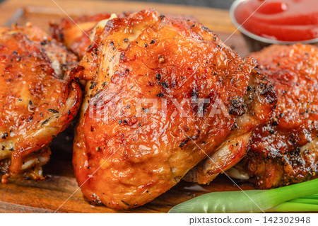 Grilled chicken thighs with crispy skin served on a wooden platter alongside a small bowl of ketchup and green onion garnish 142302948