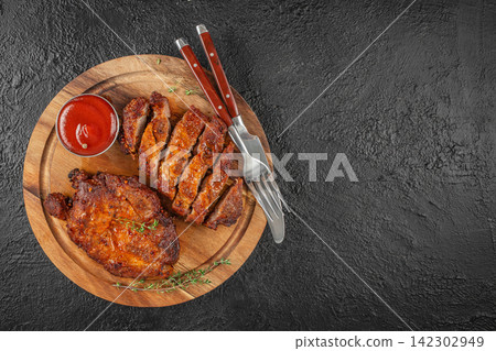 Grilled chicken breast slices arranged on a wooden plate with a small bowl of ketchup and cutlery on a textured black surface Grilled chicken breast slices arranged on a wooden plate with a small bowl of ketchup and cutlery on a textured black surface 142302949