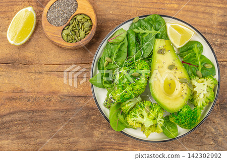Fresh green salad with avocado, broccoli, spinach, and pumpkin seeds served on a white plate with lime wedges on a wooden table Fresh green salad with avocado, broccoli, spinach, and pumpkin seeds served on a white plate with lime wedges on a wooden table 142302992
