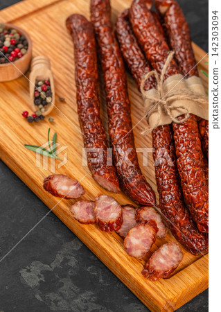 Smoked sausage links arranged on a wooden cutting board with sliced pieces, accompanied by a small bowl of mixed peppercorns and fresh herbs 142303094