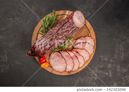 Sliced cured meat arranged on a wooden platter with whole sausage, cherry tomatoes, and fresh rosemary on a dark textured surface Sliced cured meat arranged on a wooden platter with whole sausage, cherry tomatoes, and fresh rosemary on a dark textured surface 142303096