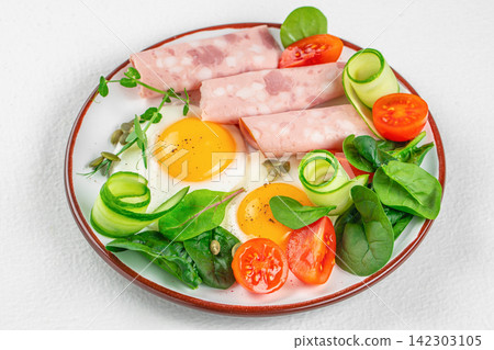 Plate of cooked eggs with slices of ham, cherry tomatoes, cucumber spirals, and fresh spinach arranged on a white background for a healthy meal presentation Plate of cooked eggs with slices of ham, cherry tomatoes, cucumber spirals, and fresh spinach arranged on a white background for a healthy meal presentation 142303105