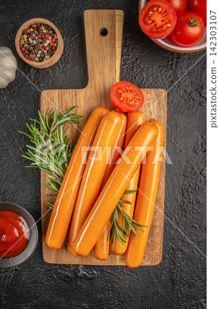 Fresh sausages arranged on a wooden cutting board with rosemary, cherry tomatoes, garlic, and spices in bowls on a textured black surface Fresh sausages arranged on a wooden cutting board with rosemary, cherry tomatoes, garlic, and spices in bowls on a textured black surface 142303107