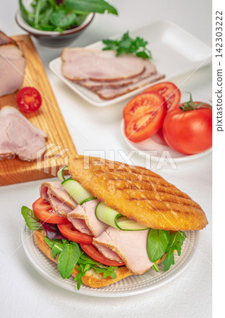 Sandwich filled with sliced turkey, fresh tomatoes, cucumbers, and leafy greens on a white plate, with additional ingredients displayed in the background Sandwich filled with sliced turkey, fresh tomatoes, cucumbers, and leafy greens on a white plate, with additional ingredients displayed in the background 142303222