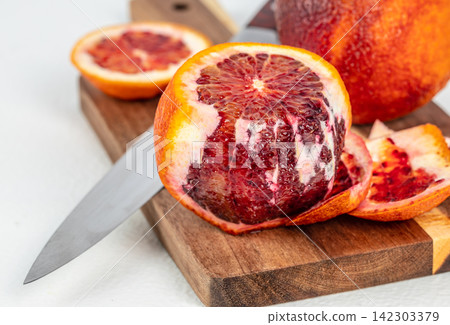 Blood orange cut in half on a wooden cutting board with a knife, showcasing vibrant red flesh and peels, with additional whole blood oranges in the background Blood orange cut in half on a wooden cutting board with a knife, showcasing vibrant red flesh and peels, with additional whole blood oranges in the background 142303379