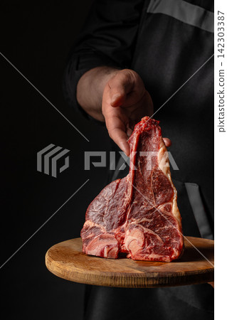 Chef in black apron presenting raw tomahawk steak on wooden cutting board against dark background, highlighting marbled texture and quality of meat Chef in black apron presenting raw tomahawk steak on wooden cutting board against dark background, highlighting marbled texture and quality of meat 142303387