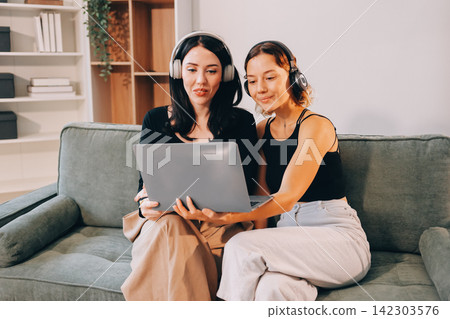 Two young female students using a laptop while sitting on a sofa at home. Student life concept with studying, teamwork, and online learning. Two young female students using a laptop while sitting on a sofa at home. Student life concept with studying, teamwork, and online learning. 142303576