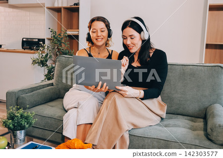 Two young female students using a laptop while sitting on a sofa at home. Student life concept with studying, teamwork, and online learning. Two young female students using a laptop while sitting on a sofa at home. Student life concept with studying, teamwork, and online learning. 142303577