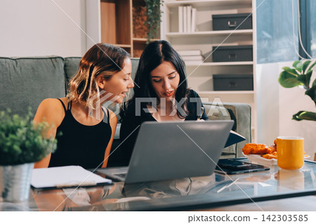 Two young female students using a laptop while sitting on a sofa at home. Student life concept with studying, teamwork, and online learning. Two young female students using a laptop while sitting on a sofa at home. Student life concept with studying, teamwork, and online learning. 142303585