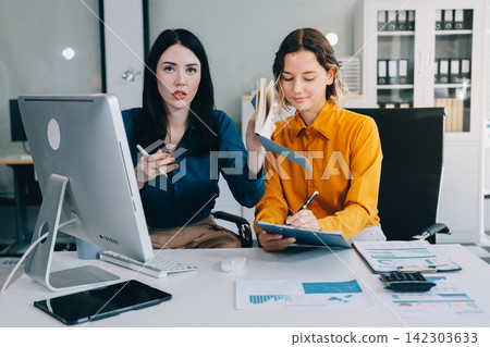 Two businesswomen discussing work using a laptop in a modern office lounge area, showing teamwork and communication Two businesswomen discussing work using a laptop in a modern office lounge area, showing teamwork and communication 142303633