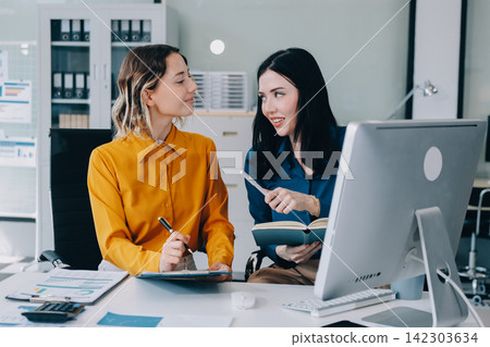 Two businesswomen discussing work using a laptop in a modern office lounge area, showing teamwork and communication Two businesswomen discussing work using a laptop in a modern office lounge area, showing teamwork and communication 142303634