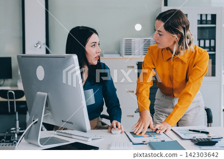 Two businesswomen discussing work using a laptop in a modern office lounge area, showing teamwork and communication Two businesswomen discussing work using a laptop in a modern office lounge area, showing teamwork and communication 142303642