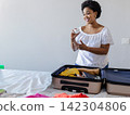 African American woman packing suitcase by bed holding white jar and showing yellow top, copy space 142304806