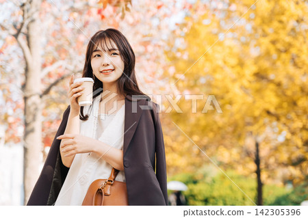 一位女子手捧一杯咖啡，漫步在遍佈美麗銀杏樹的小鎮上。 142305396