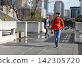 Two males walking urban walkway in red hoodie and headphones, holding phone and backpack by puddles. Cityscape, pedestrians, skyscrapers, sidewalk, planters, reflections, sunlight 142305720