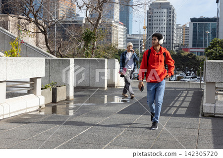 Two males walking urban walkway in red hoodie and headphones, holding phone and backpack by puddles. Cityscape, pedestrians, skyscrapers, sidewalk, planters, reflections, sunlight Two males walking urban walkway in red hoodie and headphones, holding phone and backpack by puddles. Cityscape, pedestrians, skyscrapers, sidewalk, planters, reflections, sunlight 142305720