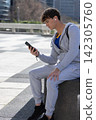 Teen male sitting on concrete seat in plaza holding phone with earbuds gray tracksuit, copy space 142305760